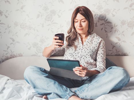 Woman sits on bed with tablet PC and cup of hot coffee. She watches online TV series. Online video call or conference, distance learning, remote education. Self isolation during quarantine.