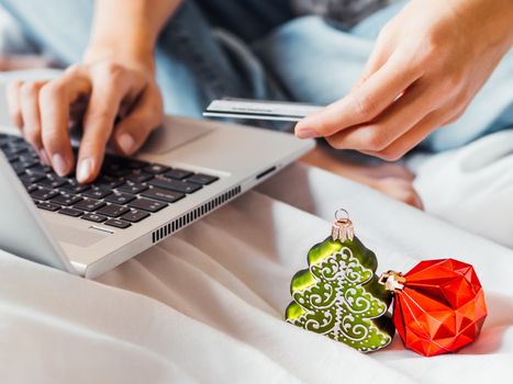 Woman using laptop to pay for online order by credit card. Decorative Christmas tree and New Year red ball in her hand. Online shopping before winter holidays.