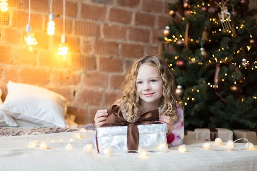 Cute Girl in pajamas lies on bed with a gift box and new year tree and lights behind