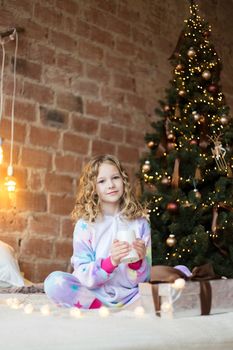 Adorable girl in pajamas sits on the bed and opens a gift against the background of New Year's garlands