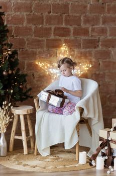 Christmas, New Year. Cute little girl in pajamas sitting on the chair at home with gift box and new year tree and lights behind