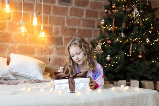 Cute Girl in pajamas lies on bed with a gift box and new year tree and lights behind