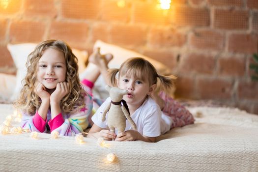 Little sisters in pajamas on the bed smiling against the background of christmas garlands
