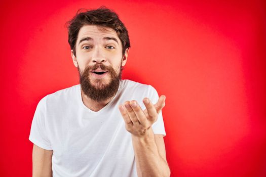 A man in a white T-shirt with a beard gestures with his hands emotions red background. High quality photo