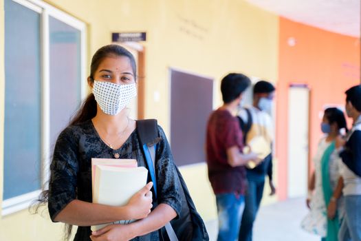 Young girl in medical mask with backpack and books standing confidently by looking into camera with students in background - concept of college reopn with safety measures due to coronavirus covid-19
