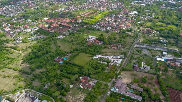 Many villas with brown-orange shingle roofs between tropical trees on the sky background in Ubud on Bali. Sun is shining onto them. Aerial photo.