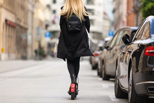 Rear view of girl riding public rental electric scooter in urban city environment at fall. New eco-friendly modern public city transport in Ljubljana.