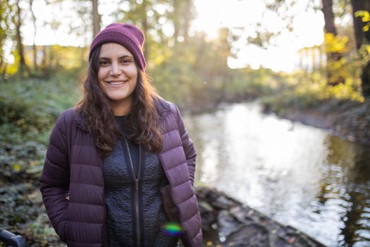 Happy brunette woman smiling and standing next to a river in the forest. Portrait of woman in forest with river as background. Adventurous day in nature