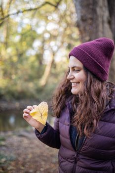 Happy woman smiling and holding an autumn leaf in the forest. Portrait of woman wearing knitted hat in forest. Adventurous day in nature