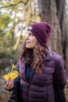 Smiling brunette woman holding a yellow leaf and looking away in the forest. Portrait of woman wearing knitted hat in forest. Adventurous day in nature