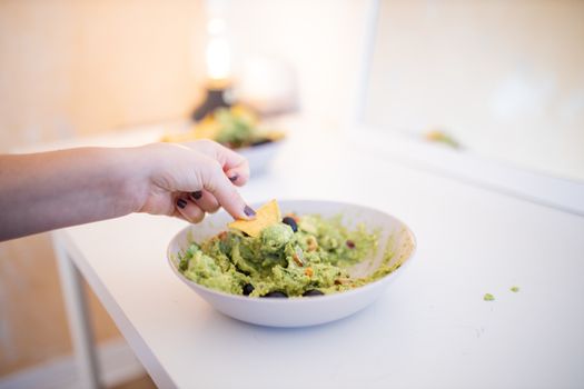 Female hand dipping nachos in guacamole sauce with olives on a white table. Bowl of traditional Mexican guacamole on a table. Spicy Mexican cuisine