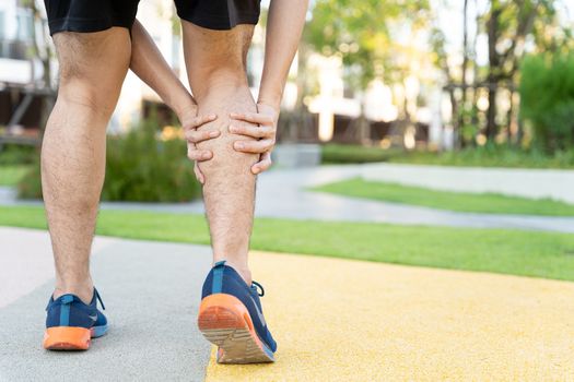 Male runner athlete leg injury and pain. Hands grab painful leg while running in the park.