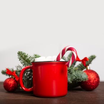 Cocoa in red mug with marshmallows candy cane fir tree branches and red baubles on wooden background with gray copy space for text