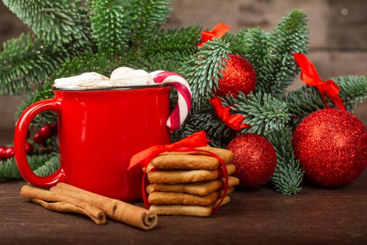 Cocoa hot chocolate in red mug with marshmallows candy cane gingerbread cookies fir tree branches and red baubles on dark wooden background