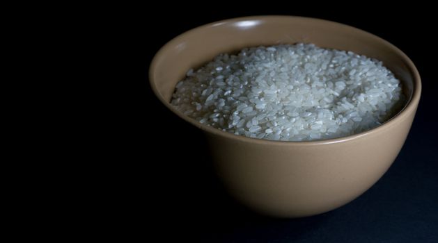 White uncooked rice in a beige bowl with scattered grains nearby on a black background