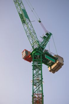Vertical sunrise image looking up at a tall green and orange tower crane. Full frame crane and cabin with a plain blue sky background.
