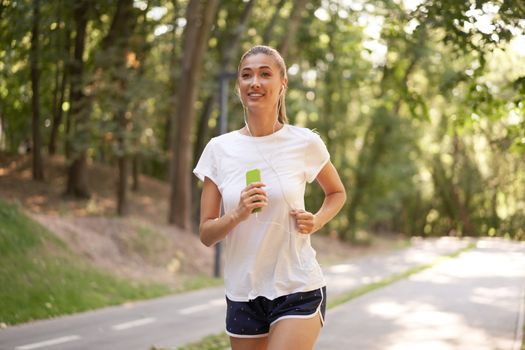 Woman listen music running on asphalt track summer park Caucasian female earphones jogging sunny morning Runner training hold smartphone in hand Healthy lifestyle positive concept
