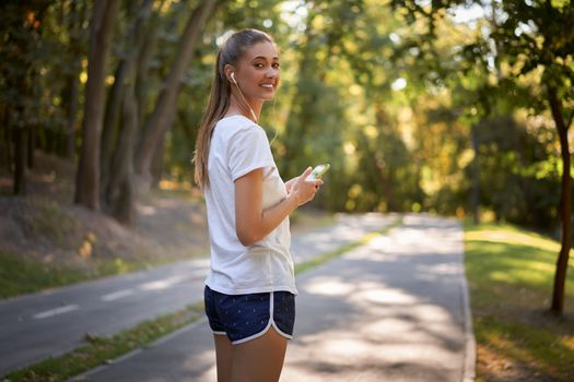 Woman putting on earphones to listen music before jogging summer park Caucasian female preparing to run sunny morning Runner ready training holdin smartphone hand Healthy lifestyle positive concept