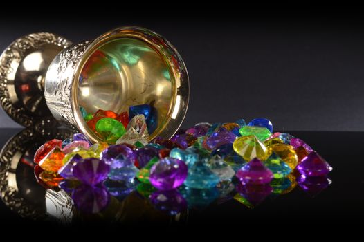 A closeup view of some treasured gemstones and a silver wineglass over a black reflective background.