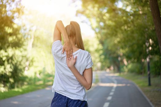 Woman runner stretching arms before exercising summer park morning Middle age athletic female warming up body before running Caucasian person warm up jogging Dressed white shirt shorts running track