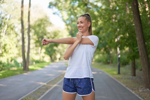 Woman runner stretching arms before exercising summer park morning Middle age athletic female warming up body before running Caucasian person warm up jogging Dressed white shirt shorts running track