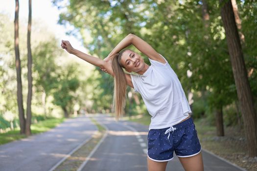 Woman runner stretching arms before exercising summer park morning Middle age athletic female warming up body before running Caucasian person warm up jogging Dressed white shirt shorts running track