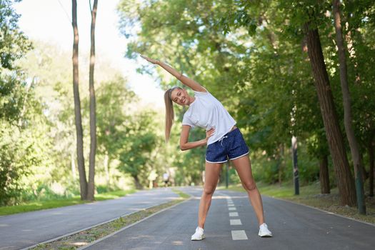 Woman runner stretching arms before exercising summer park morning Middle age athletic female warming up body before running Caucasian person warm up jogging Dressed white shirt shorts running track