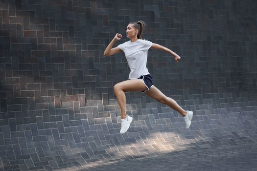 Woman with fit body jumping and running against black wall background. Female model in sportswear exercising outdoors. Sport motivation Healthy lifestyle concept full length