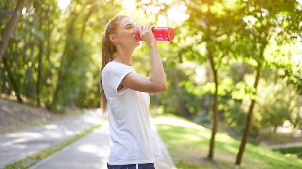 Woman drink water red bottle after morning workout. Young athletic female rear view walk asphalt running track park after jogging Healthy lifestyle concept