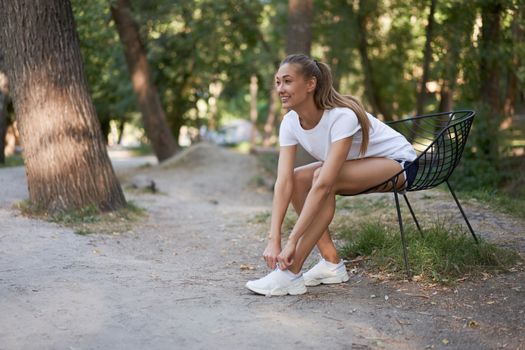 Woman sitting chair tie shoelaces on sneakers after morning workout. Young athletic female rear view walk asphalt running track park after jogging Healthy lifestyle concept