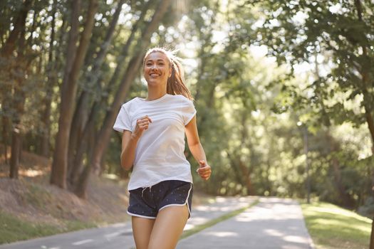 Woman running asphalt road summer park Active sporty caucasian female morning workout Healthy lifestyle concept. Athletic person sportswear exercises Dressed white t-shirt