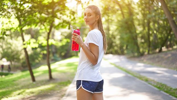 Woman drink water red bottle after morning workout. Young athletic female rear view walk asphalt running track park after jogging Healthy lifestyle concept