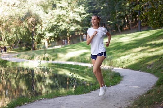 Woman running road summer park near lake Active sporty caucasian female morning workout Healthy lifestyle concept. Athletic person sportswear exercises Dressed white t-shirt full length