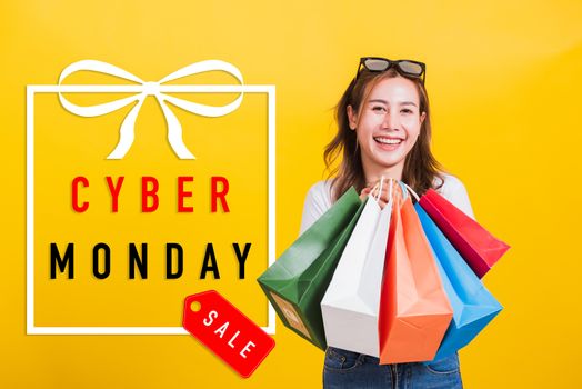 Asian happy portrait beautiful young woman smiling stand with sunglasses excited holding shopping bags multi color looking camera with Cyber Monday text in gift box on side isolated yellow background