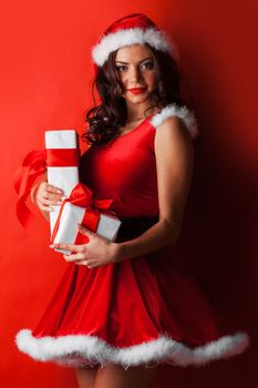 Beautiful young woman in red Santa Claus style clothes holding Christmas gifts, red background