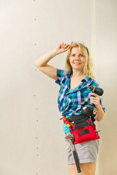 Blond woman wearing a DIY tool belt full of a variety of tools on a unpainted plasterboard wall background. DIY woman concept.