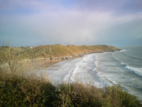 Approaching high-tide at Caswell Bay Beach in Wales, UK. Stunning view of crashing waves onto the sandy shores from the Gower Coastal Path towards sunset.