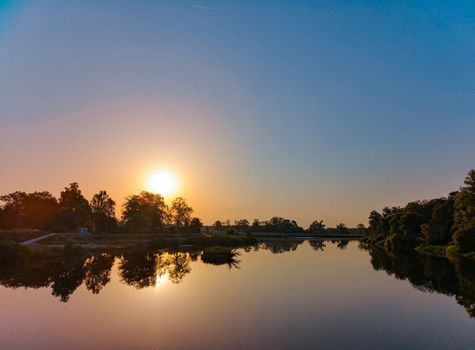 Rising sun over long river with waves between trees at sunny morning