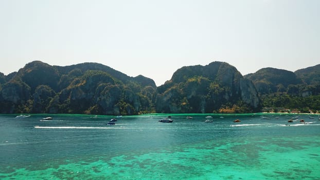 Turquoise clear water. Yachts, boats floating. Water Gradient from light to dark blue. Phi Phi don island, shooting from a drone from the air. White sand, green trees, palm trees and large hills.