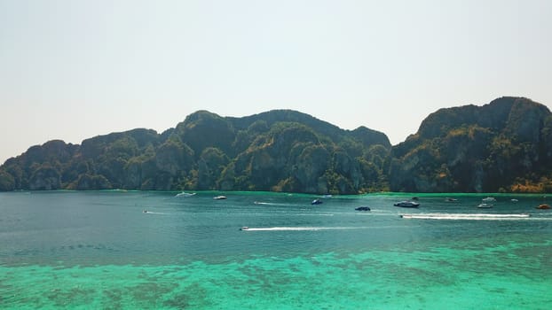 Turquoise clear water. Yachts, boats floating. Water Gradient from light to dark blue. Phi Phi don island, shooting from a drone from the air. White sand, green trees, palm trees and large hills.