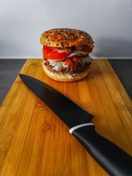 Hamburger with roll with sesame tomato bacon cheese at end of wooden board with black chef knife