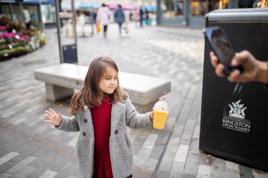 Happy little girl confidently holding a yellow smoothie while she is recorded with a smartphone. Young child with cold fruit beverage. Cute kids enjoying drinks