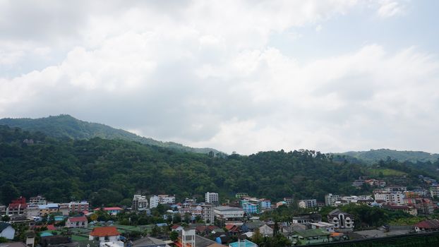 Drone view of the city of Patong, Phuket island. Houses of different heights stand on the beach. Green hills of the island. On the roof of houses pools, people swim. On the road go scooters and cars.