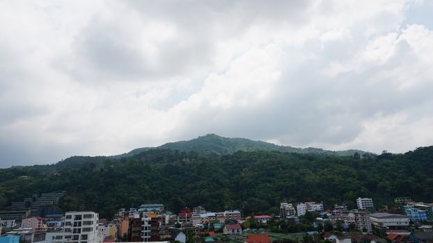 Drone view of the city of Patong, Phuket island. Houses of different heights stand on the beach. Green hills of the island. On the roof of houses pools, people swim. On the road go scooters and cars.