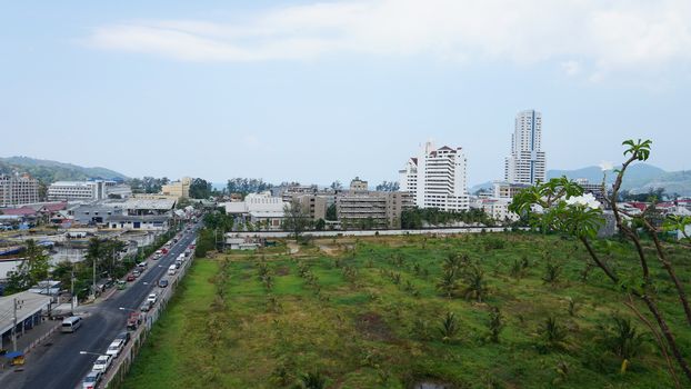 Drone view of the city of Patong, Phuket island. Houses of different heights stand on the beach. Green hills of the island. On the roof of houses pools, people swim. On the road go scooters and cars.