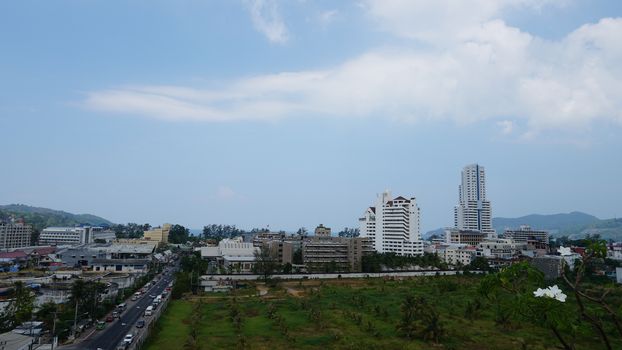 Drone view of the city of Patong, Phuket island. Houses of different heights stand on the beach. Green hills of the island. On the roof of houses pools, people swim. On the road go scooters and cars.