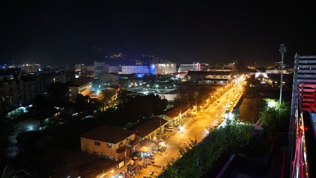 Night city of Patong, Phuket island. Houses of different heights stand on the beach. Street lights are shining brightly. On the road go scooters and cars. Tropical climates, green palm trees.