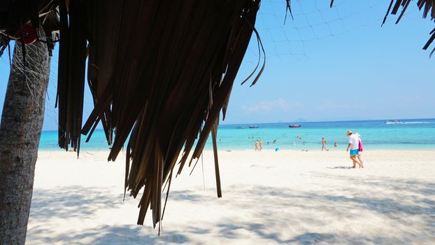 Palm leaves on the roof of the gazebo. Beach with white sand and blue water. People are actively spending time. Blue sky with clouds. The trunk of the palm tree. Bamboo Island, Thailand.