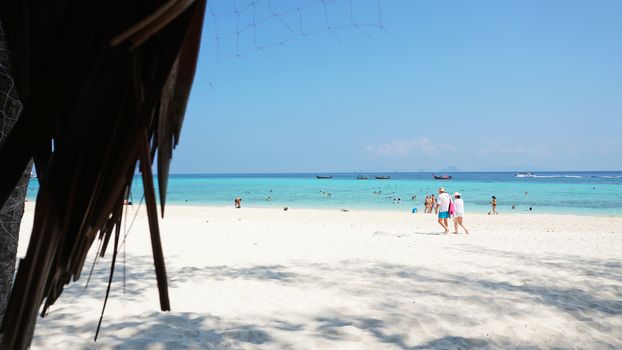 Palm leaves on the roof of the gazebo. Beach with white sand and blue water. People are actively spending time. Blue sky with clouds. The trunk of the palm tree. Bamboo Island, Thailand.