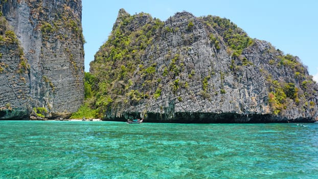 Blue water, green hills and steep rocks. The boat sails near the island. Boat with tourists in the Bay. Steep cliffs and caves. Group of tourists on the boat. Sea excursion. Water gradient.